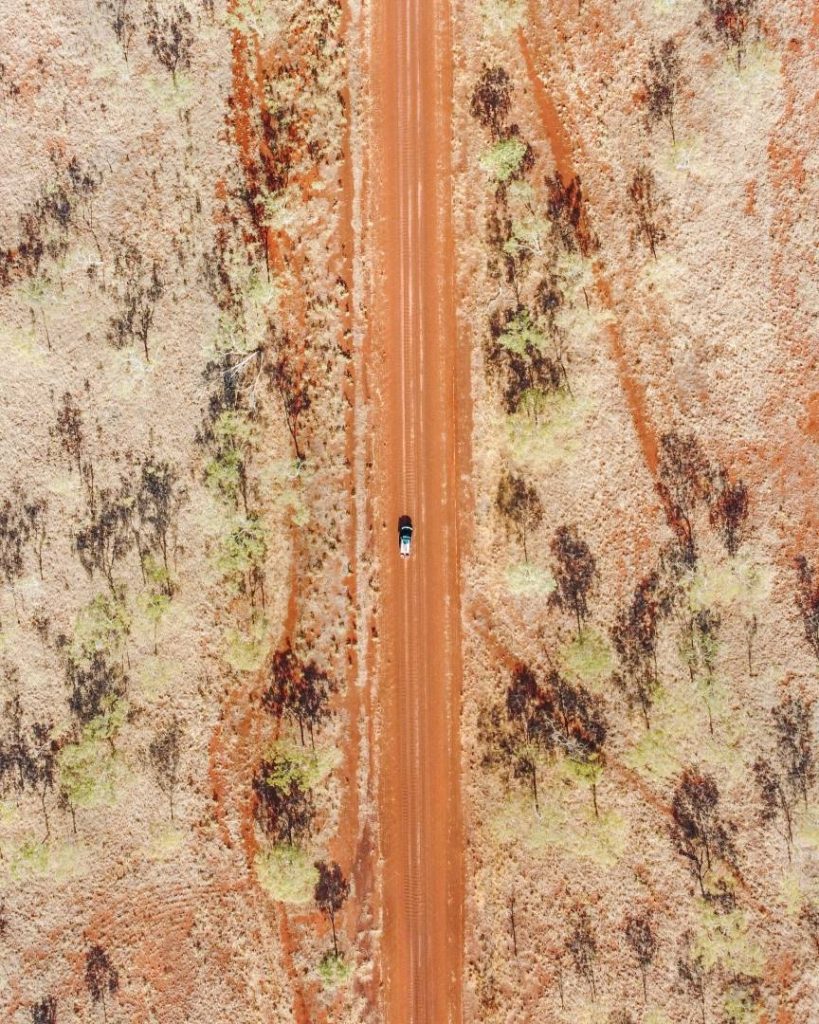 Aerial view of the Gibb River Road cutting through red dirt terrain with sparse vegetation, featuring a single vehicle on the road.