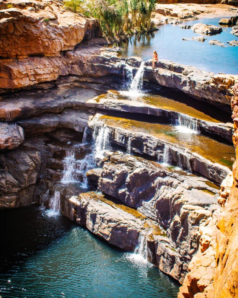 A person sitting at the edge of a tiered waterfall in Bell Gorge, surrounded by rugged cliffs and clear pools, in Gibb River Road, Western Australia