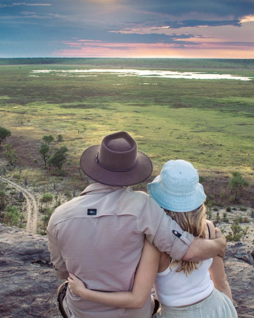 A couple sitting on Ubirr Rock, watching the sun set over the wetlands of Kakadu National Park, with vibrant skies in the distance.
