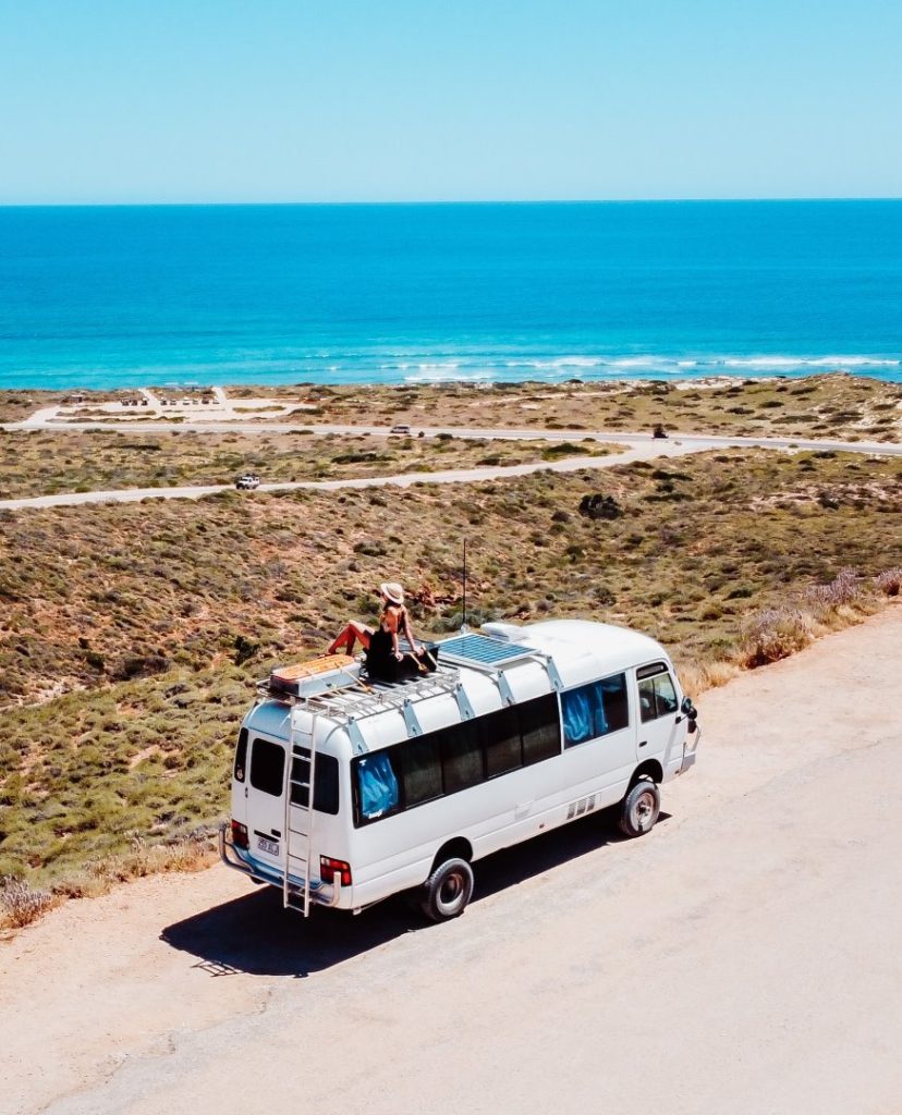A white camper van parked near the coastline in Exmouth, with a traveler sitting on the roof overlooking the ocean