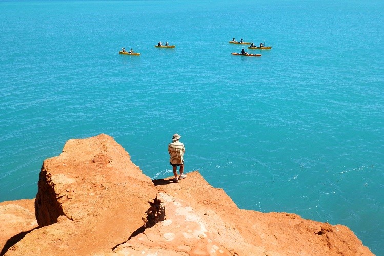 A person standing on red cliffs overlooking kayakers paddling in the calm blue waters near Broome.