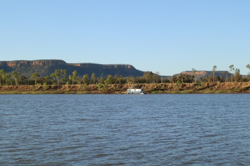 A peaceful view of the Victoria River with a houseboat on the water and rugged cliffs in the distance.
