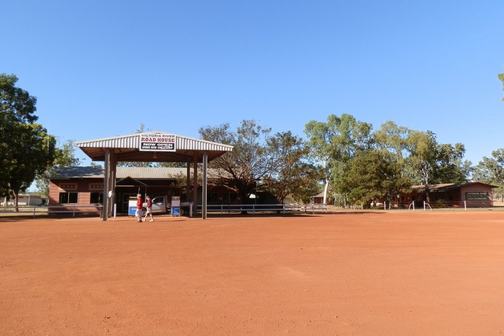 The Victoria River Roadhouse, featuring a rustic building with a sign reading "Victoria River Roadhouse" against a backdrop of red dirt and trees.