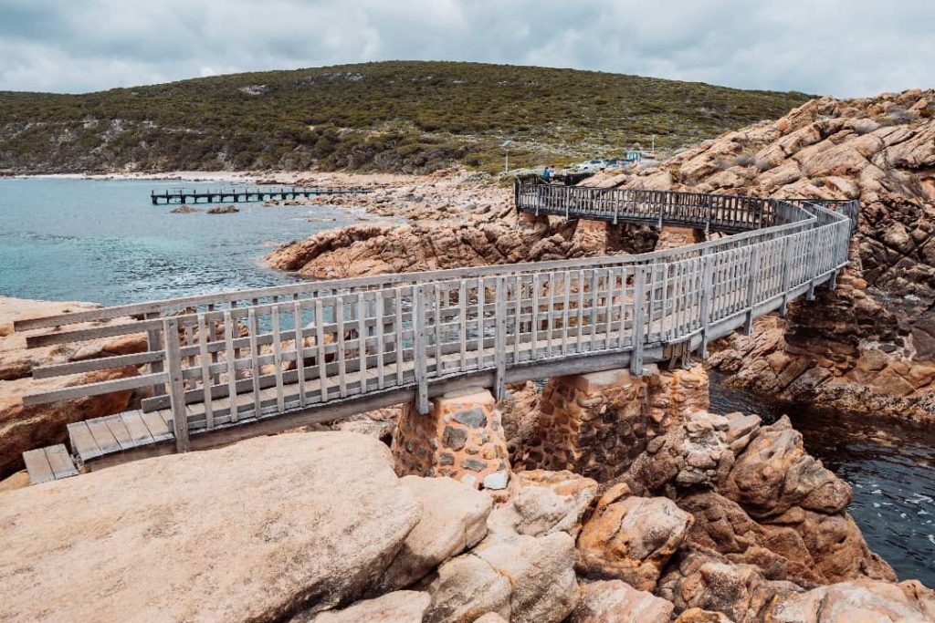 A wooden boardwalk winding through rugged rock formations at Canal Rocks, with turquoise ocean waters and a distant pier in the background.