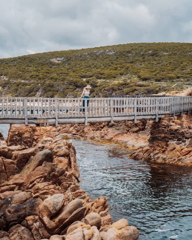 A woman in a wide-brim hat walking on a wooden boardwalk above a rocky canal at Canal Rocks, with greenery-covered hills in the background.