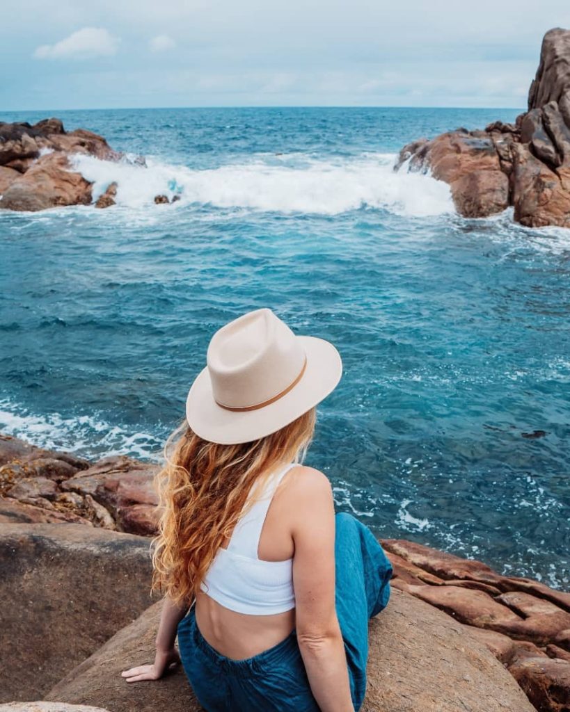 A woman with long hair and a beige hat sitting on a rock, gazing at the waves crashing against the rugged rock formations at Canal Rocks.