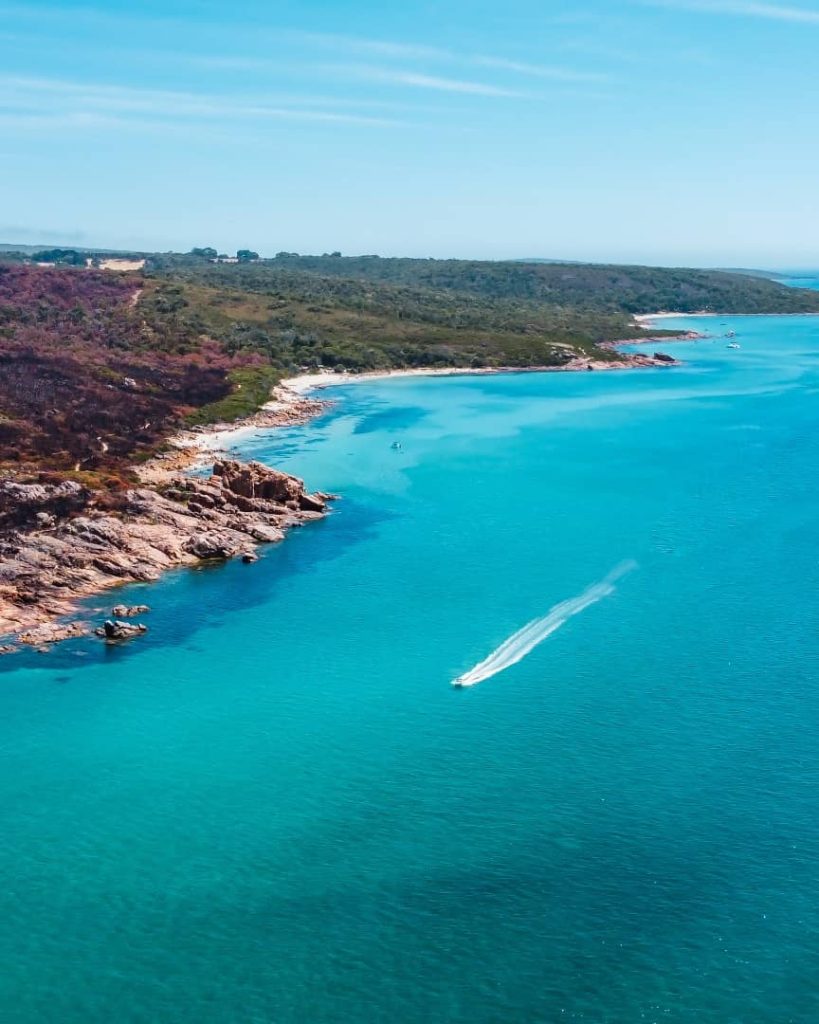 A speedboat glides across turquoise waters near the rocky coastline of Castle Rock, Dunsborough, Western Australia, with lush greenery and a clear blue sky in the background.