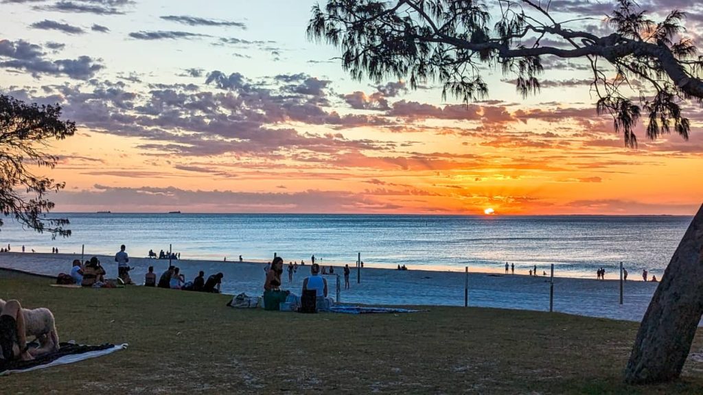 A vibrant sunset over City Beach, Perth, Western Australia, with people relaxing on the grass and strolling along the shoreline as the sun dips below the horizon.