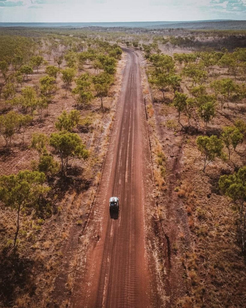 A vehicle travels down a red dirt track on the Gibb River Road, surrounded by sparse bushland and open skies in the remote Australian outback.
