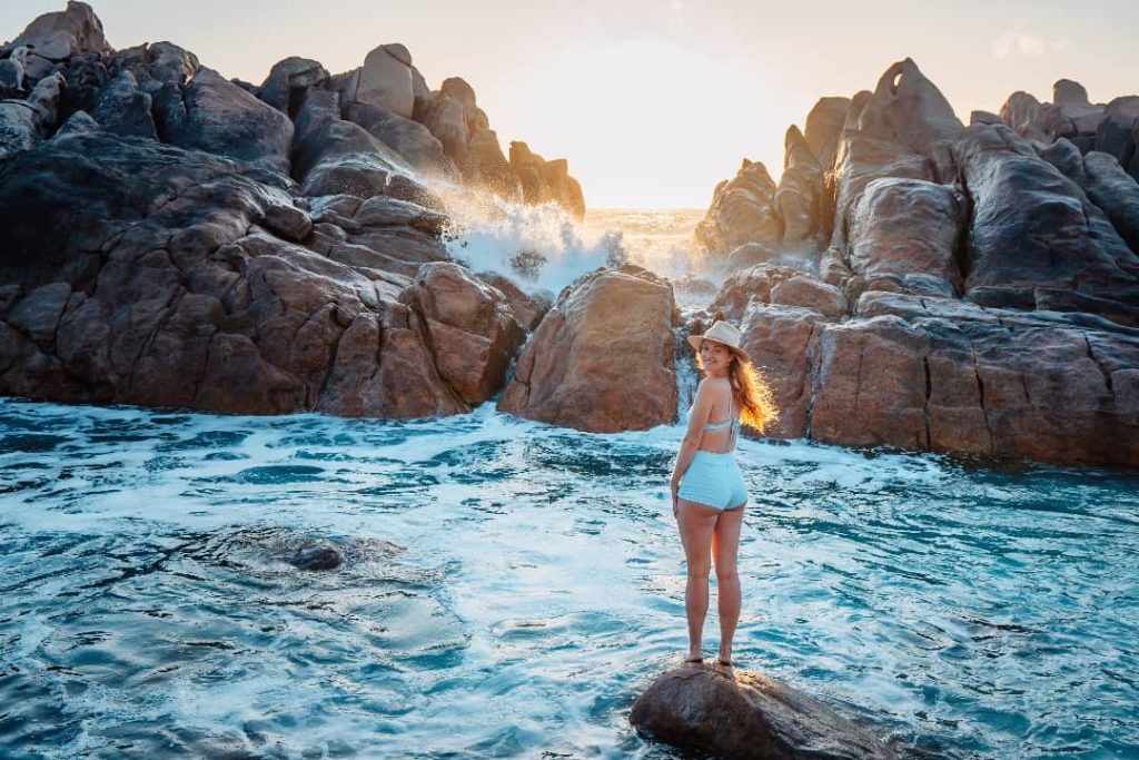 A woman in a blue swimsuit and sun hat stands on a rock at Injidup Natural Spa, Western Australia, with waves crashing against rugged cliffs at sunset.