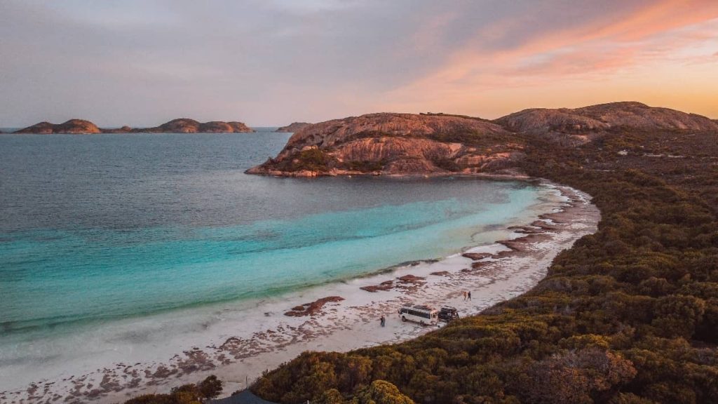 A scenic view of Lucky Bay, Esperance at sunset, with turquoise water meeting white sand and rugged hills in the background. A few vehicles and people are visible along the beach.