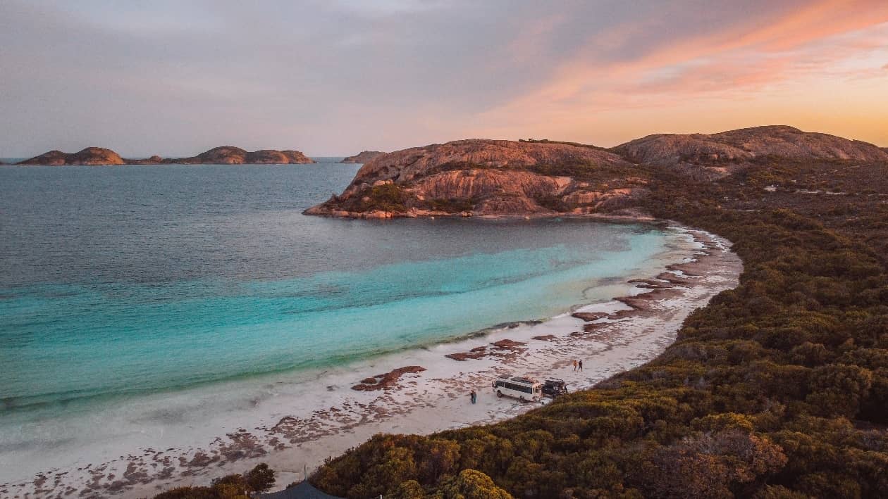 A scenic view of Lucky Bay, Esperance at sunset, with turquoise water meeting white sand and rugged hills in the background. A few vehicles and people are visible along the beach.
