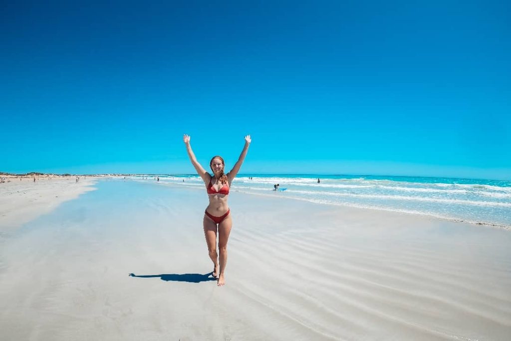 A woman in a red bikini stands on the sandy shore of Cable Beach, Broome, with her arms raised joyfully under a bright blue sky.