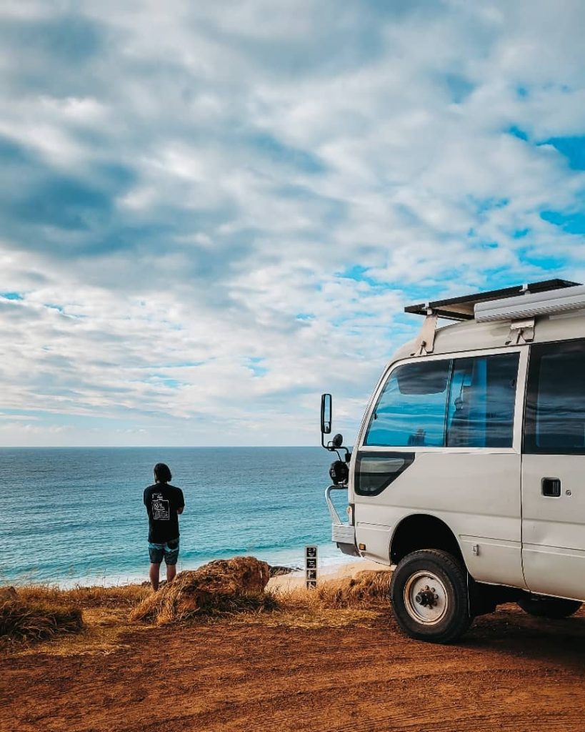 A man stands at a coastal lookout, gazing at the ocean under a partly cloudy sky, with a campervan parked nearby on a dirt road.