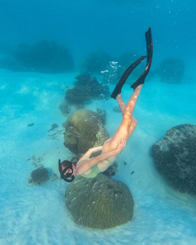  woman snorkeling underwater in clear turquoise waters, wearing fins and a mask, surrounded by large coral formations on the sandy ocean floor in Western Australia.
