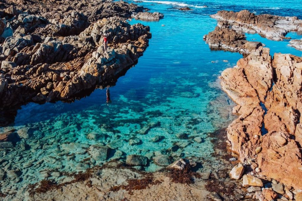 Crystal-clear blue water surrounded by rugged, reddish-brown rock formations at "The Aquarium" in Yallingup, with a person standing on the rocks admiring the view.