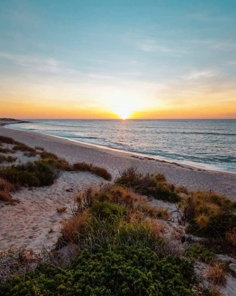 A peaceful sunset over Wobiri Beach in Exmouth, Western Australia, with golden hues reflecting off the ocean and sandy dunes in the foreground.
