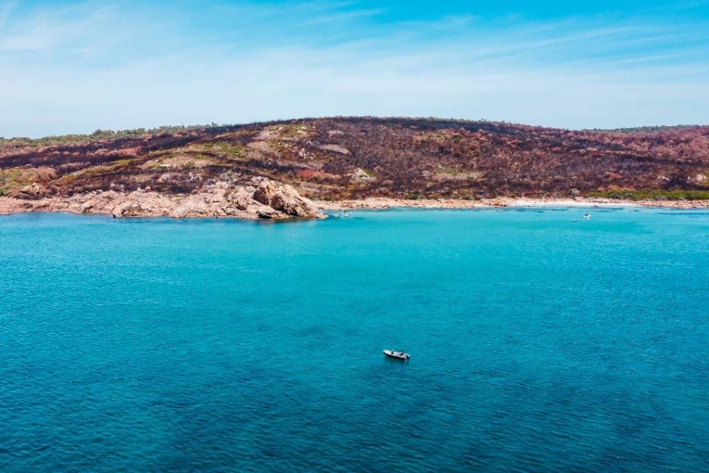 A small boat floating in calm, turquoise waters near Yallingup, Western Australia, with a rocky shoreline and rugged hills in the background under a clear blue sky.