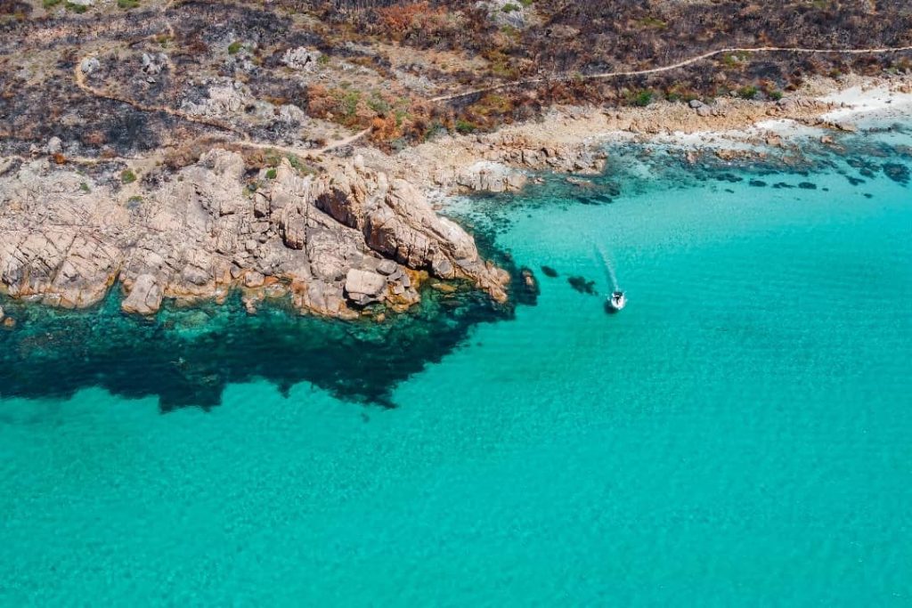 A small boat cruising through crystal-clear turquoise waters near rocky cliffs in Yallingup, Western Australia. The surrounding landscape features burnt vegetation from a previous fire, contrasting with the vibrant coastal scenery.