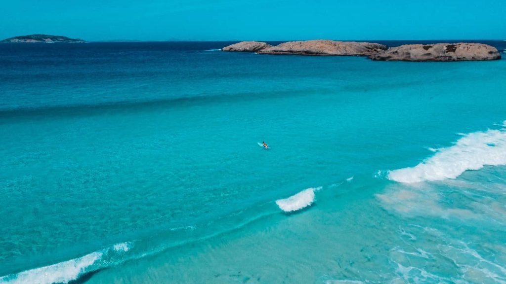 A person paddles through the crystal-clear turquoise waters of Twilight Beach in Esperance, Western Australia, with gentle waves rolling onto the shore and rocky islands in the distance.