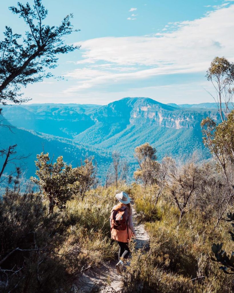 A hiker on a narrow trail surrounded by native vegetation, overlooking the expansive Blue Mountains terrain. Spring is the best time to visit Blue Mountains for hiking, with crisp air and vibrant foliage adding charm to the landscape.
