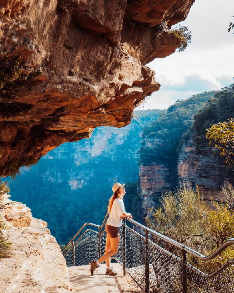 A hiker pauses on a narrow cliffside path, overlooking the forested valleys of Wentworth Falls in the Blue Mountains. Winter offers cool temperatures and clear skies for breathtaking walks like this.