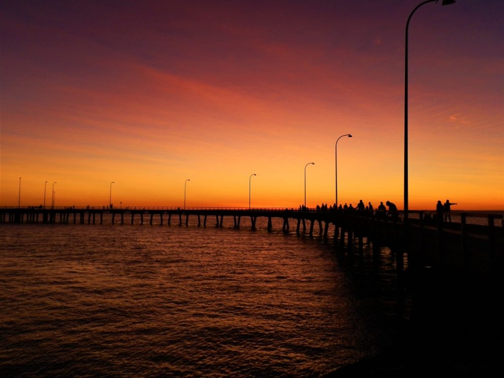 A glowing orange sunset over Derby Jetty in the Kimberley region of Western Australia, with silhouettes of people on the pier.