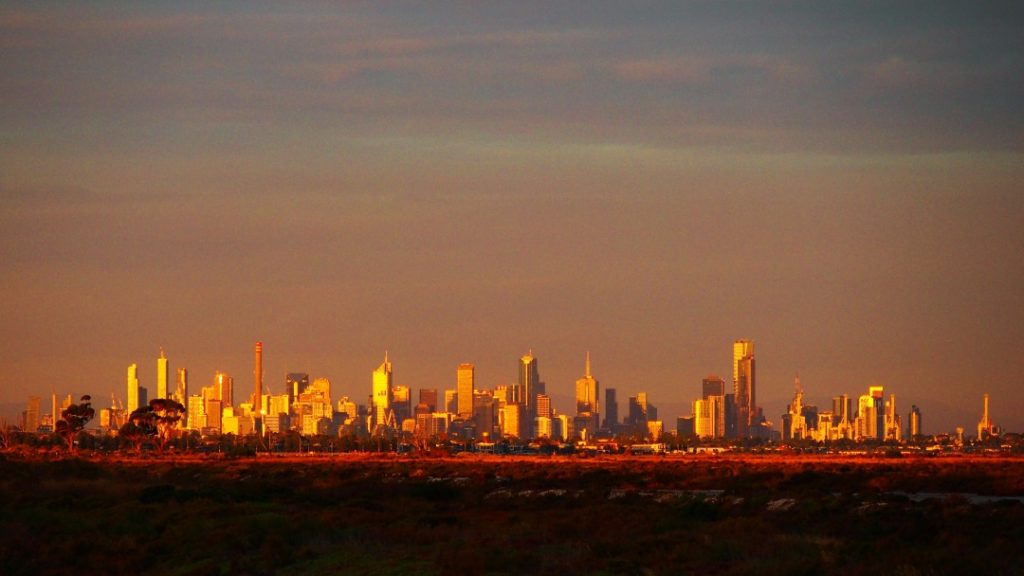 The Melbourne city skyline glows in golden hues during dusk in Victoria, Australia, with a mix of modern skyscrapers and natural landscapes in the foreground.