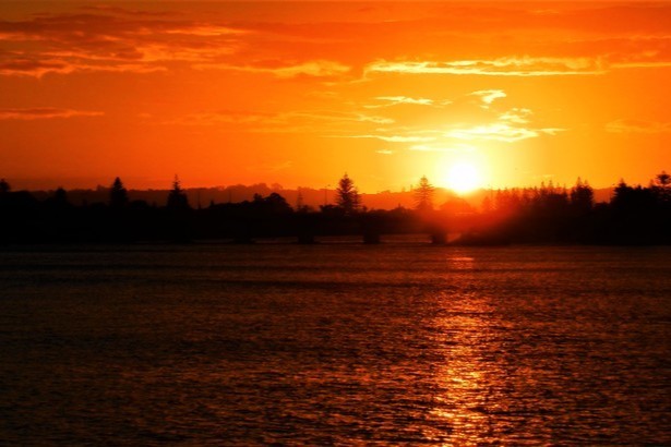 The sun sets over the Richmond River in Ballina, New South Wales, Australia, casting a warm orange hue across the water and silhouetted trees.