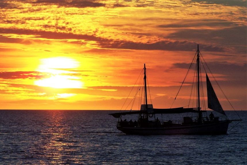 A vibrant sunset over the ocean in Darwin, Northern Territory, Australia, with a silhouetted sailboat peacefully floating on the water.
