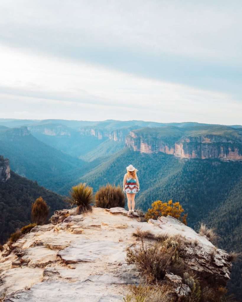 A woman standing on the edge of Burramoko Ridge with panoramic views of the Blue Mountains, a highlight of great tours in the region. Towering cliffs and endless forests create a dramatic scene.