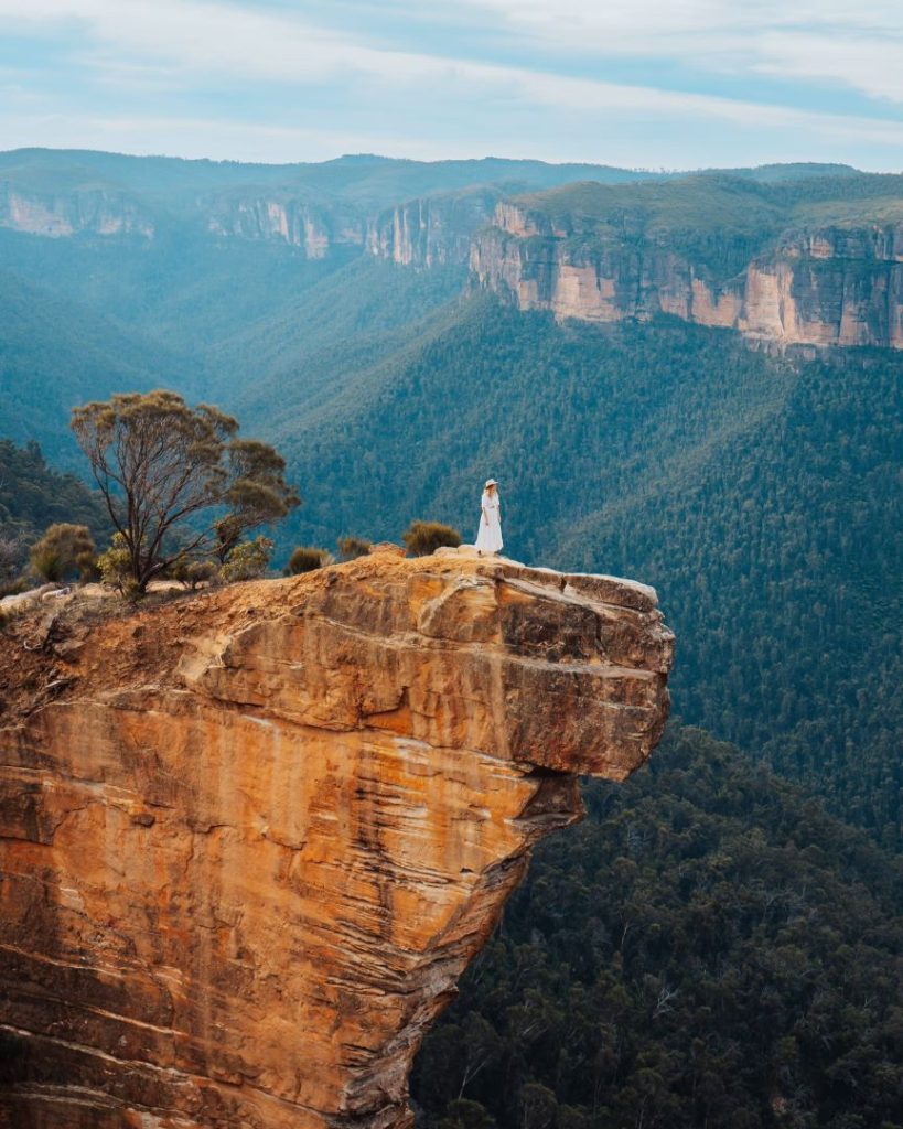 A woman with a white dress standing on the iconic Hanging Rock / Burramoko Ridge, a popular destination for the best tours in the Blue Mountains. The precarious rock formation offers jaw-dropping views.