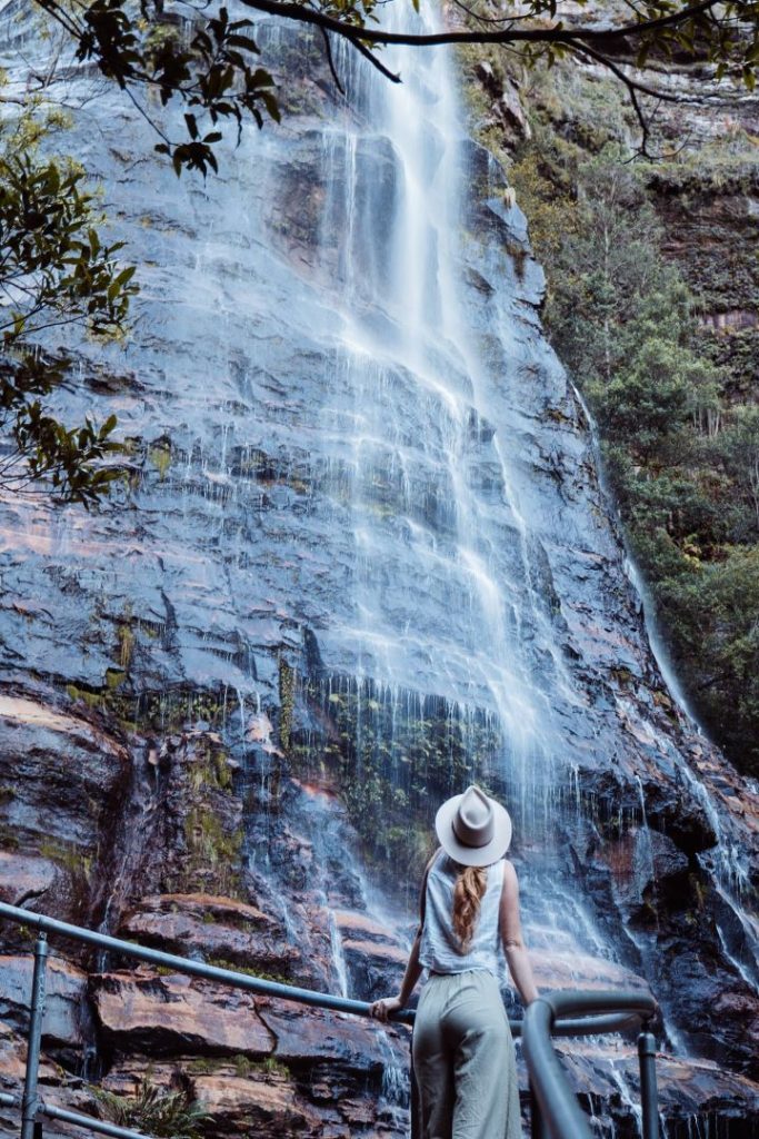 A hiker stands near a cascading waterfall at Leura Cascades, surrounded by lush foliage. Spring is the best season for this hike, as the waterfalls are at their most vibrant.