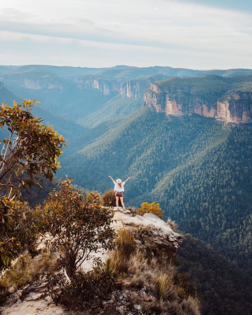 A hiker stands on the edge of Hanging Rock, overlooking the vast valleys of the Blue Mountains under a bright sky. Summer provides long daylight hours for panoramic views, but spring remains the best time for cooler hikes.
