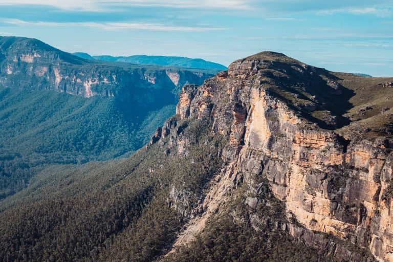 A breathtaking view of the Blue Mountains in New South Wales, Australia, with towering sandstone cliffs, lush forested valleys, and rolling hills under a clear blue sky.