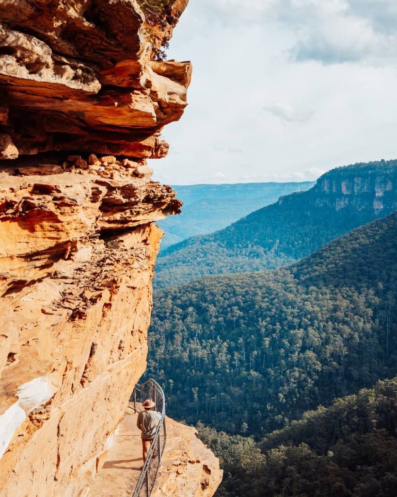 A hiker walks along a narrow cliffside path with safety railings at Wentworth Falls in the Blue Mountains, New South Wales, Australia, with sweeping views of lush forested valleys and dramatic sandstone cliffs.