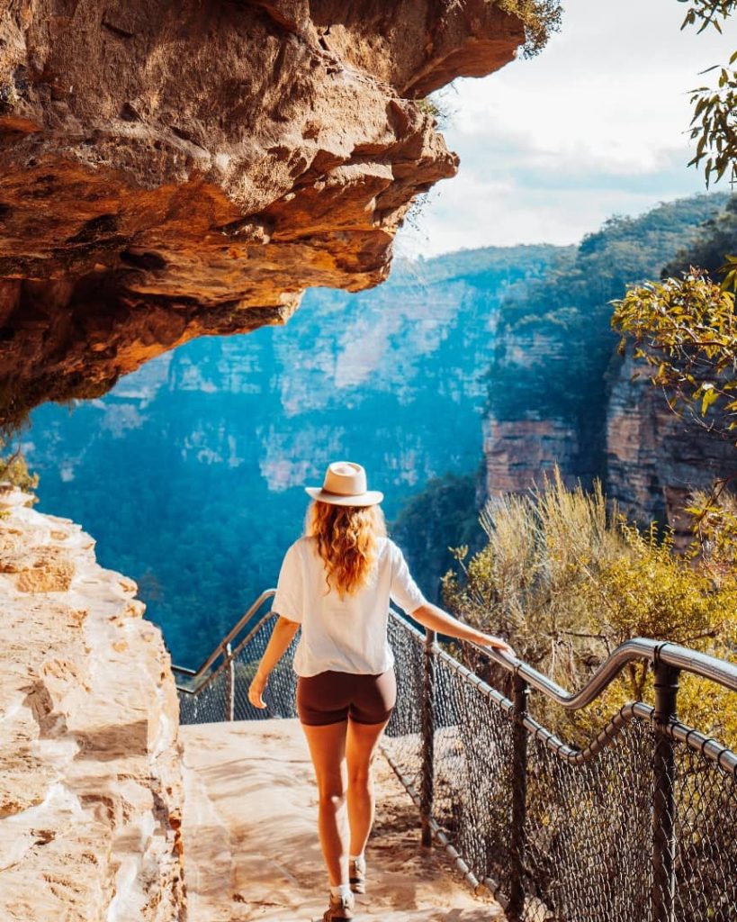 A woman in a hat walking along a scenic cliffside path at New South Wales, with breathtaking sandstone cliffs in the background.