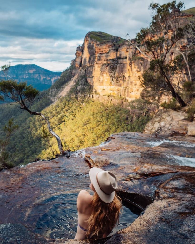 A woman with long hair and a hat sits in a natural infinity pool at Fortress Falls in the Blue Mountains, New South Wales, Australia, with dramatic sandstone cliffs and lush forest in the background.