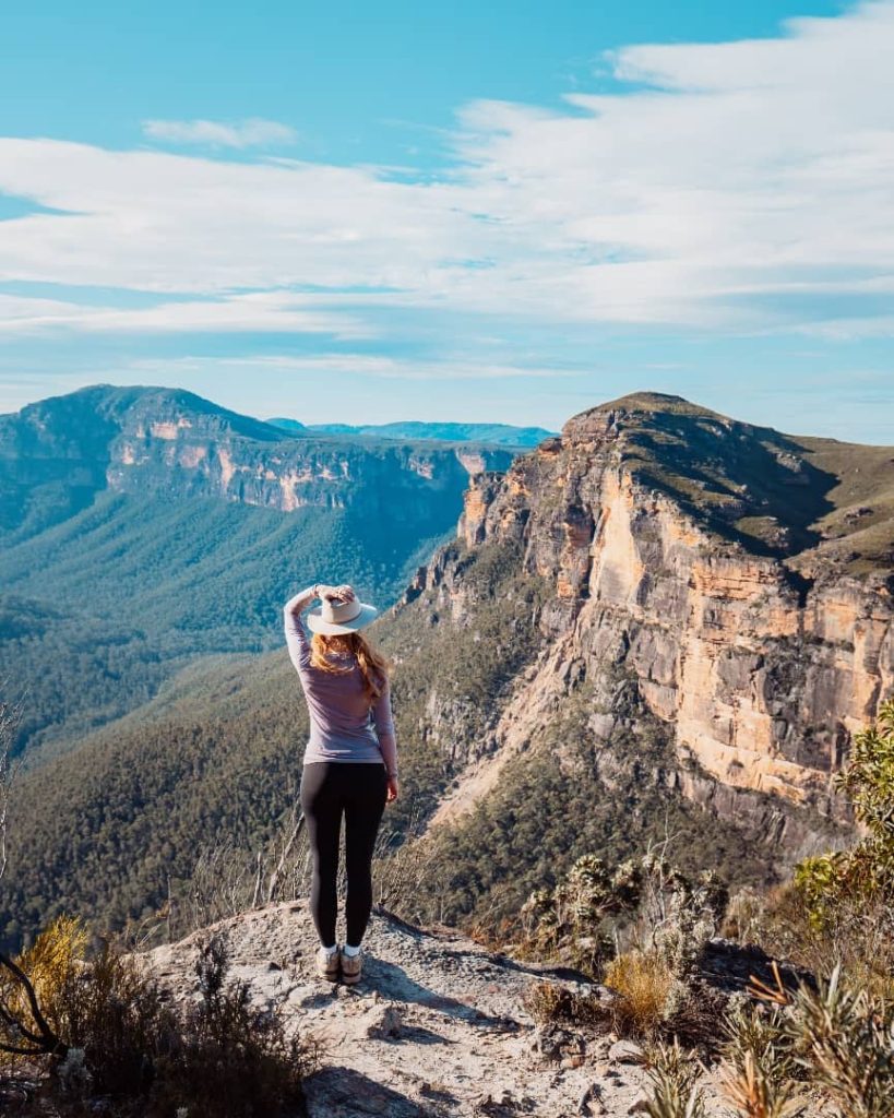 A woman in hiking gear and a hat stands on a rocky ledge overlooking the Blue Mountains in New South Wales, Australia, with vast forested valleys and sandstone cliffs stretching into the distance under a bright blue sky.