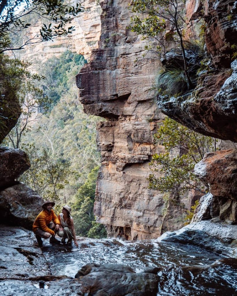 Two people wearing hats sit by a cascading stream in a secluded area of the Blue Mountains, framed by rugged sandstone cliffs and lush greenery.