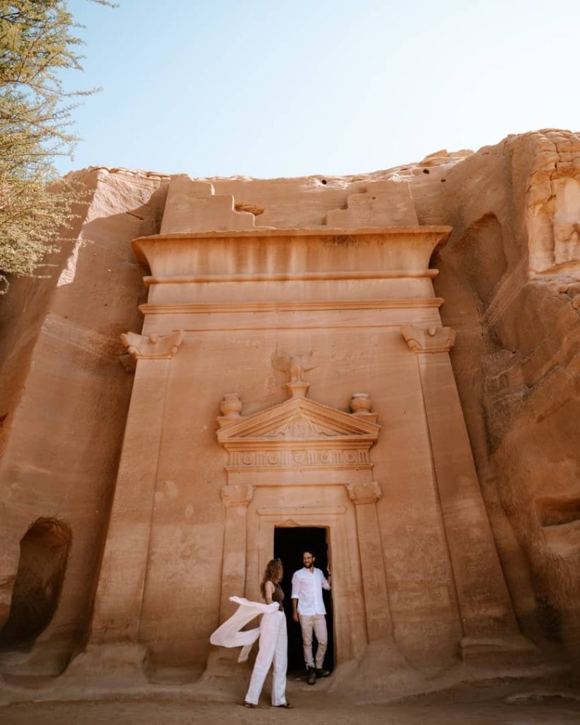 A man and woman stand at the entrance of an intricately carved Nabatean tomb in Mada'in Saleh, AlUla, Saudi Arabia. The sandstone structure showcases detailed architectural features under a bright blue sky, reflecting the area's rich historical heritage.