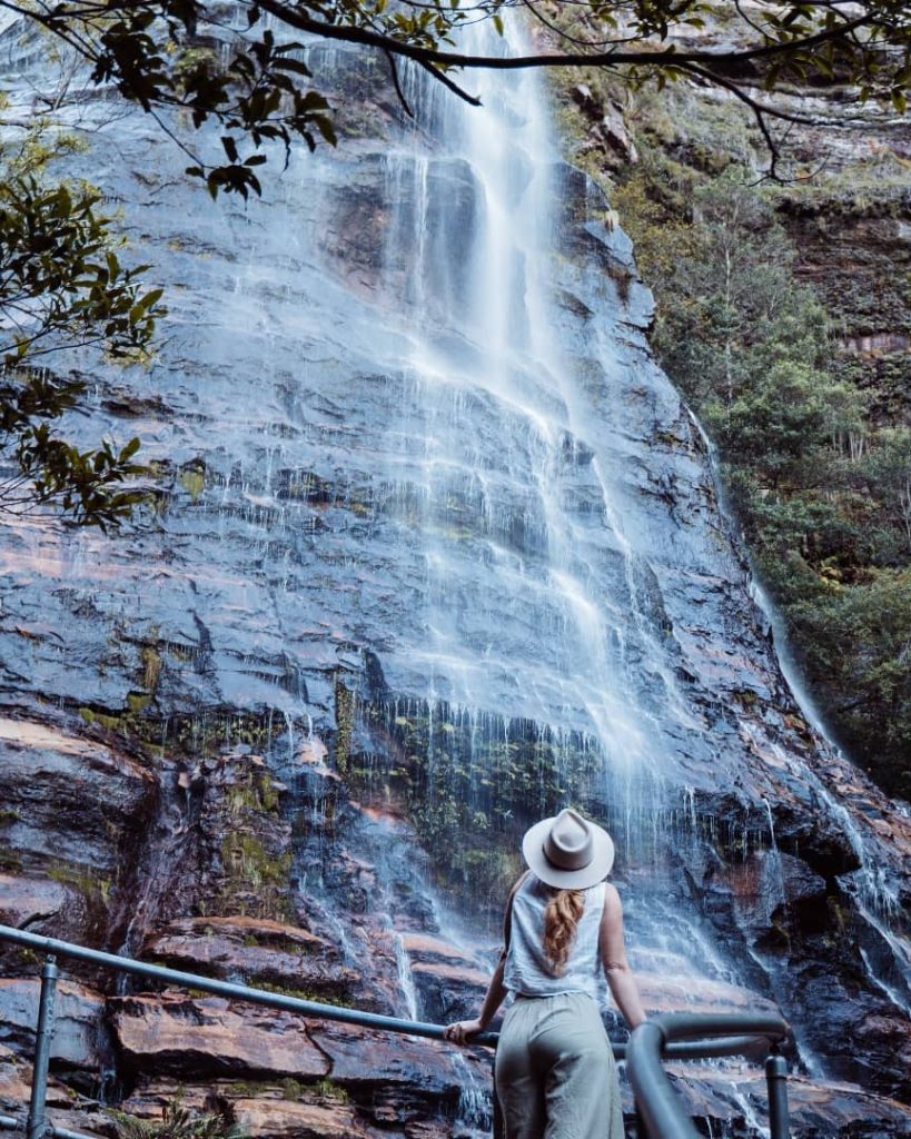 A woman wearing a hat stands on a viewing platform, gazing up at a tall cascading waterfall surrounded by lush greenery in the Blue Mountains, New South Wales, Australia.