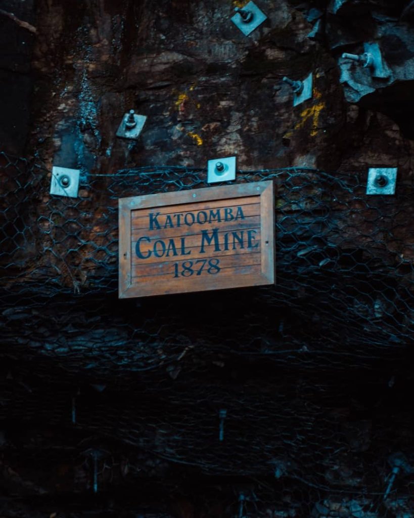 A wooden sign reading "Katoomba Coal Mine 1878" mounted on a rock face with metal support and mesh netting, located in the Blue Mountains, New South Wales, Australia.