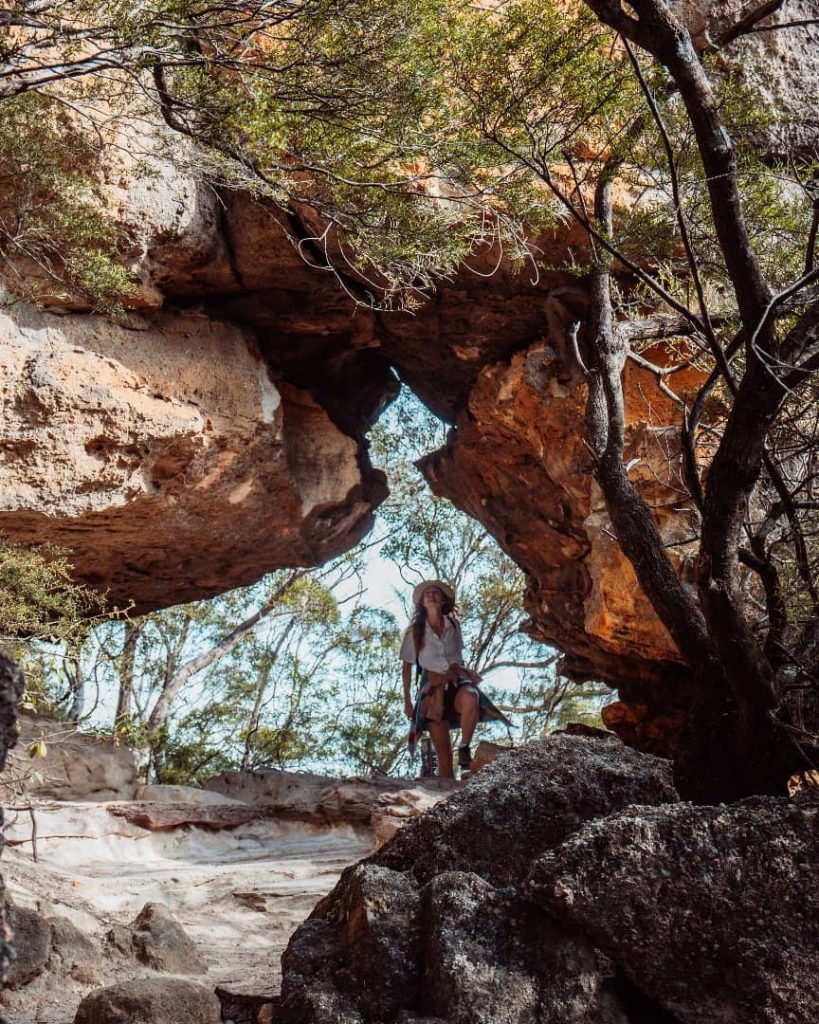 A hiker in a white shirt and hat is seated beneath a natural rock arch surrounded by trees during a hike at Wentworth Falls, Blue Mountains, New South Wales, Australia.
