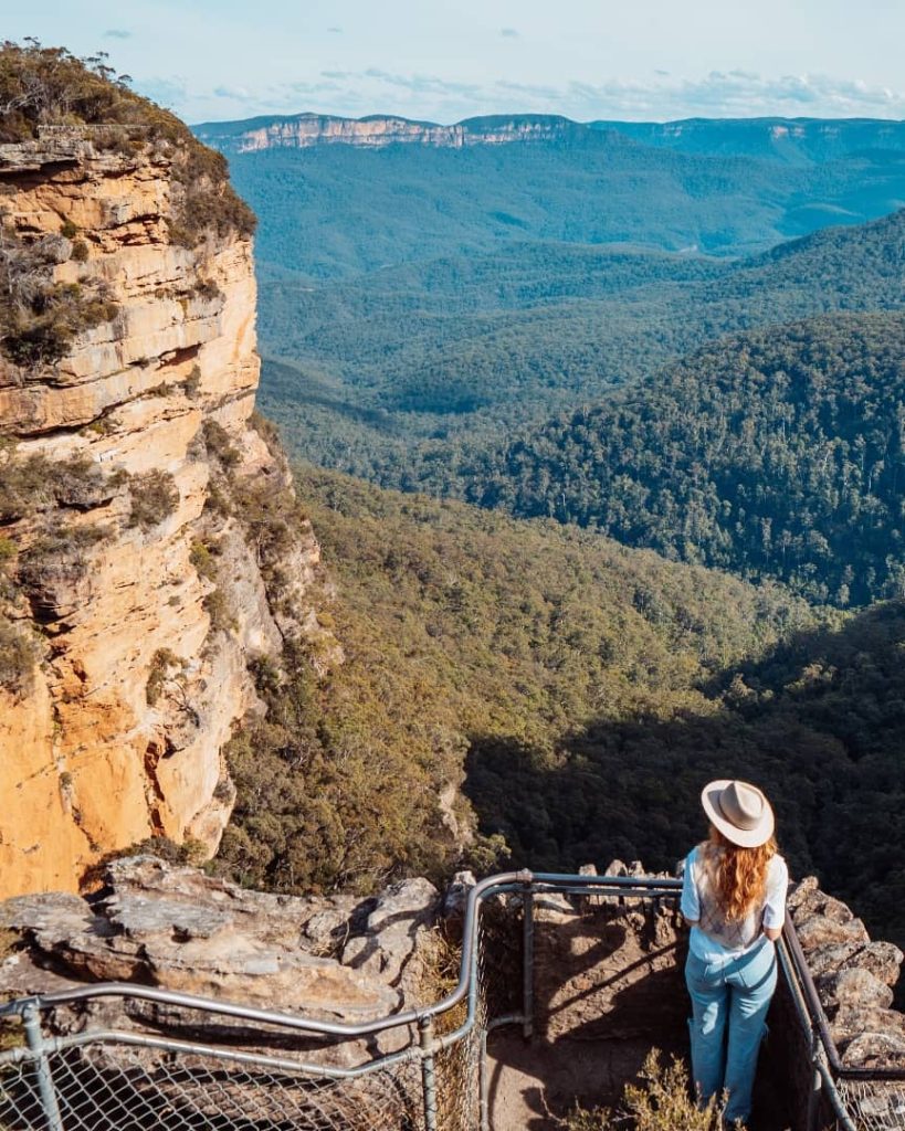 A woman wearing a white hat and light blue outfit stands at the edge of Echo Point Lookout in the Blue Mountains, NSW. She gazes over the vast valley below, where dense green forests stretch between towering sandstone cliffs. The golden sunlight highlights the rugged rock formations, enhancing the breathtaking landscape.