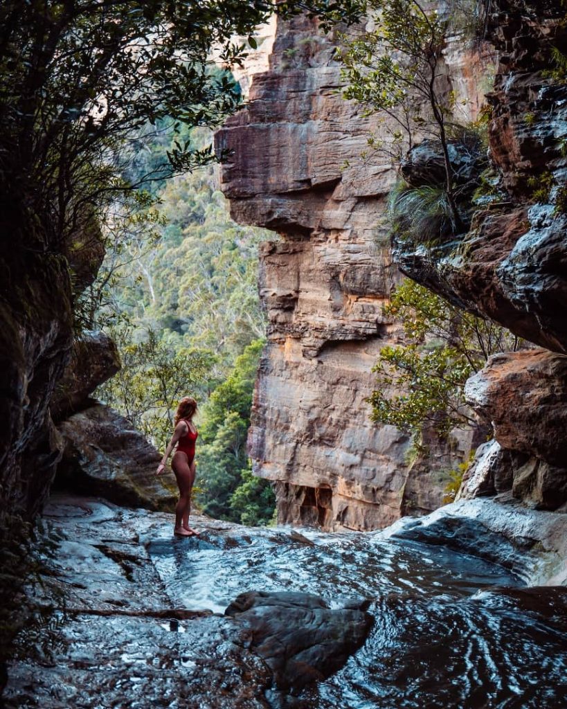 A woman in a red swimsuit stands at the edge of a natural infinity pool at Fortress Falls in the Blue Mountains, NSW. She gazes at the towering sandstone cliffs and lush green valley below as water flows gently over the rocky ledge. The secluded spot is framed by overhanging trees and moss-covered rocks, creating a peaceful and hidden oasis.