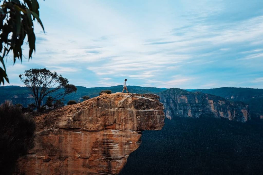 A person wearing a hat and light-colored outfit walks along the narrow edge of Hanging Rock, a dramatic sandstone cliff in the Blue Mountains, NSW. The sheer drop beneath them emphasizes the rugged beauty of the towering rock formations and dense forest in the background. The cloudy sky adds to the moody, adventurous atmosphere of the remote location.