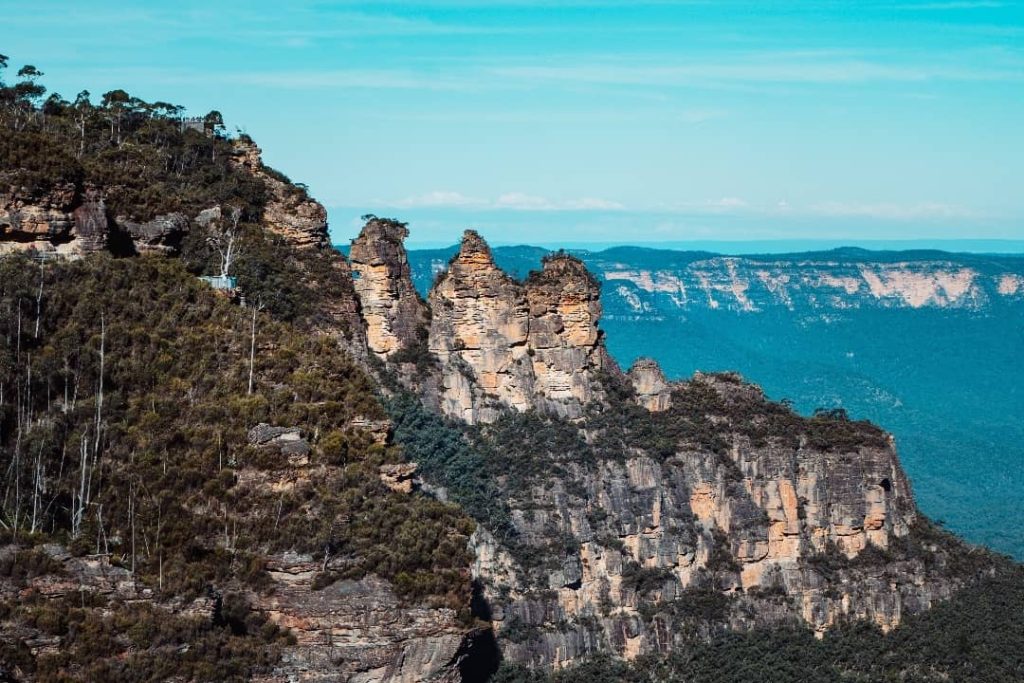 The Three Sisters, a famous rock formation in the Blue Mountains, Australia, rise sharply from a forested ridge. The three sandstone pillars stand against a backdrop of deep valleys and distant cliffs under a clear blue sky. The rugged landscape is covered in lush green vegetation, emphasizing the contrast between rock and wilderness.