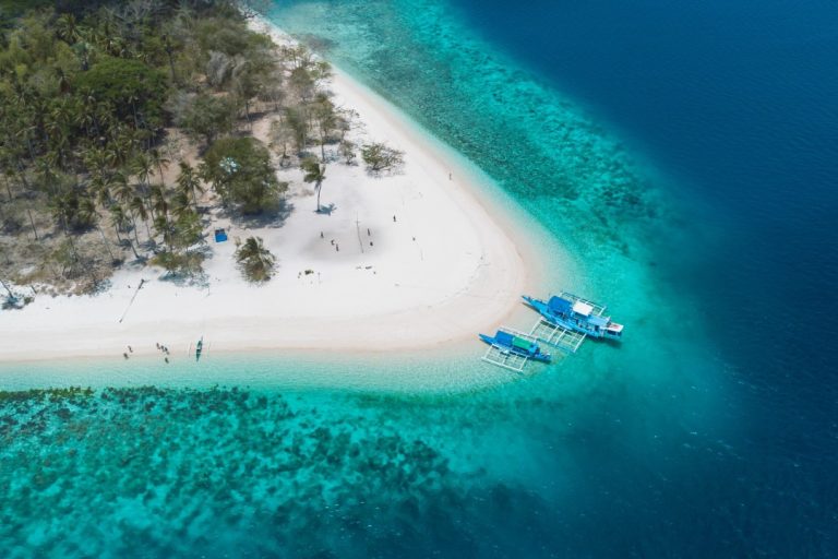 Aerial view of a tropical island in the Philippines with white sandy shores, turquoise waters, and two blue outrigger boats docked near the beach. This stunning island is one of the best places to visit in the Philippines, offering pristine beaches and crystal-clear waters.