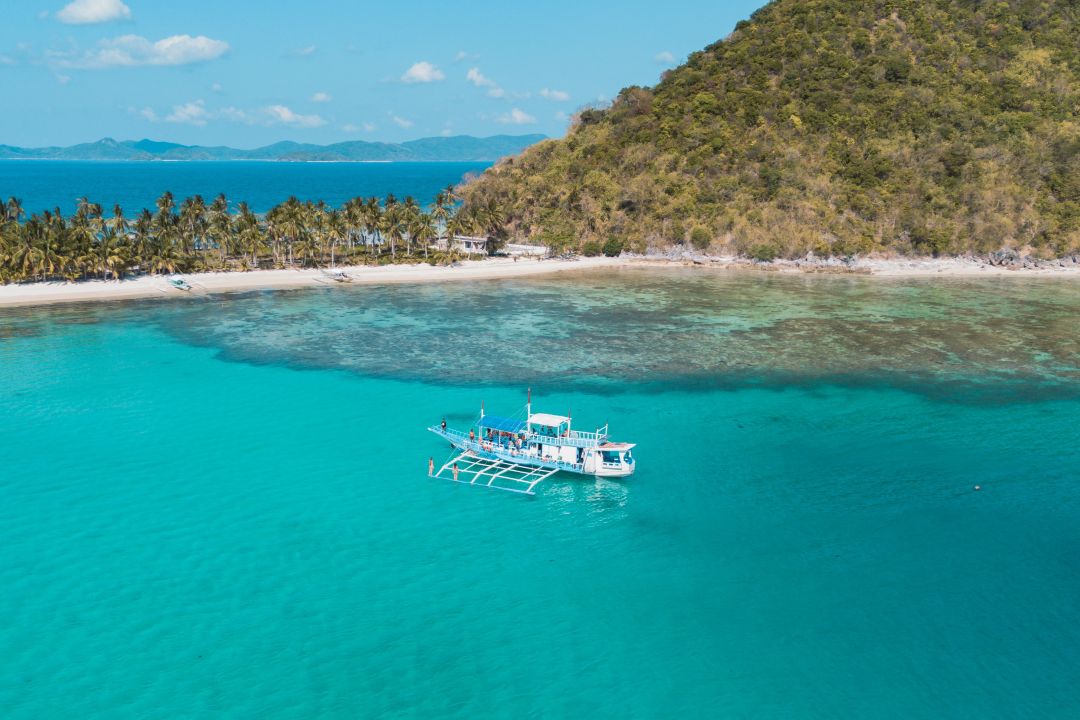 A traditional Filipino outrigger boat floats in bright turquoise waters near a palm-lined beach and lush hill in Coron, Palawan, one of the best tours in Coron for island hopping.